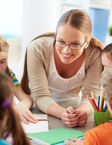 Female teacher with glasses smiling while leaning on a table with students sitting at it.