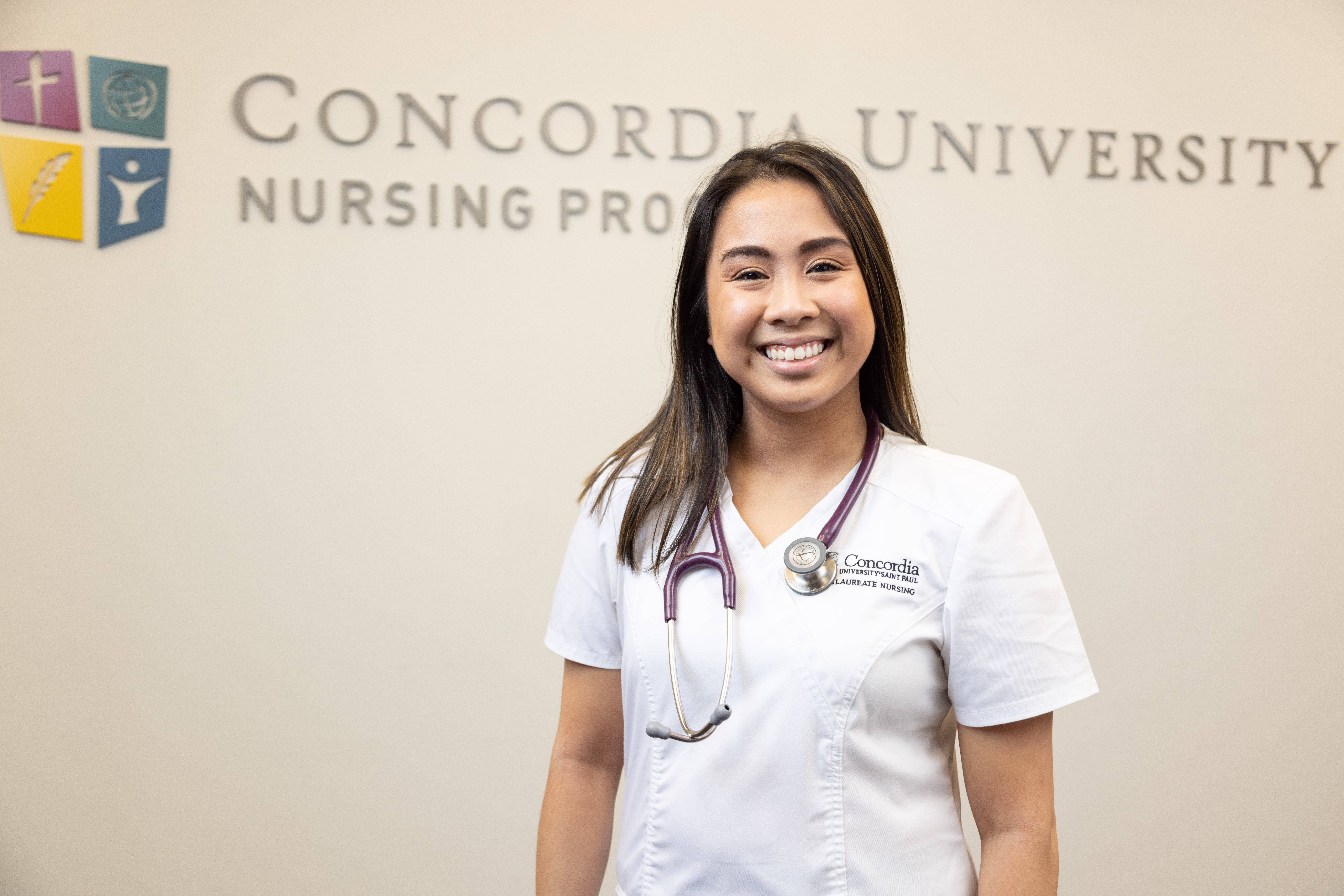 Smiling nursing student wearing white scrubs and a purple stethoscope standing in front of a tan wall that has a Concordia University Nursing Program logo on it.
