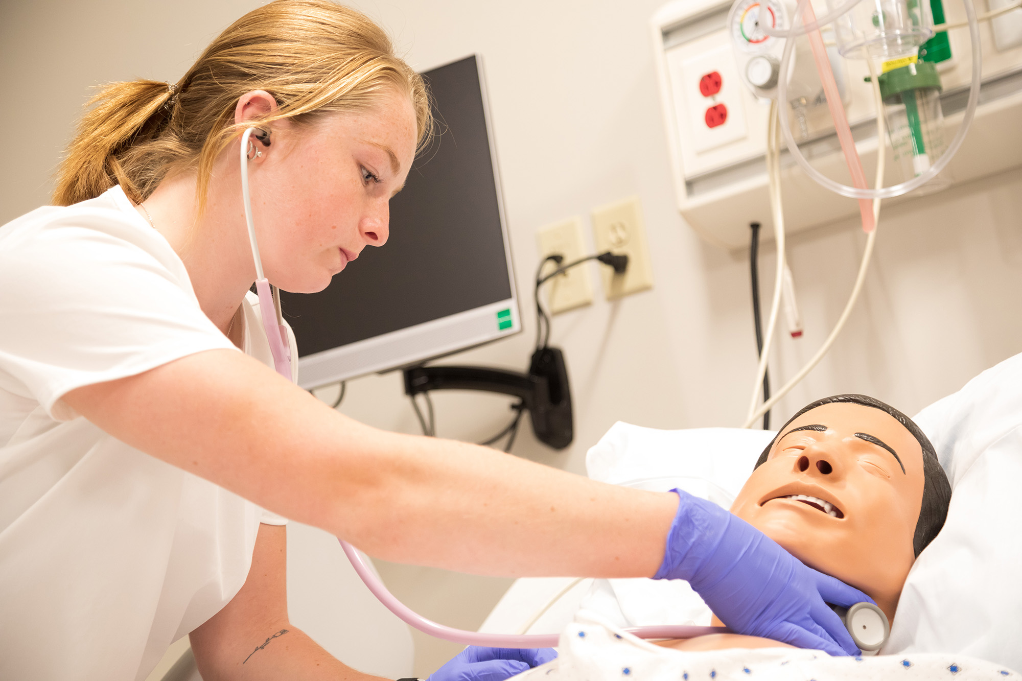 Nursing student wearing white scrubs with a pink stethoscope leaning over a learning dummy performing clinical learning.