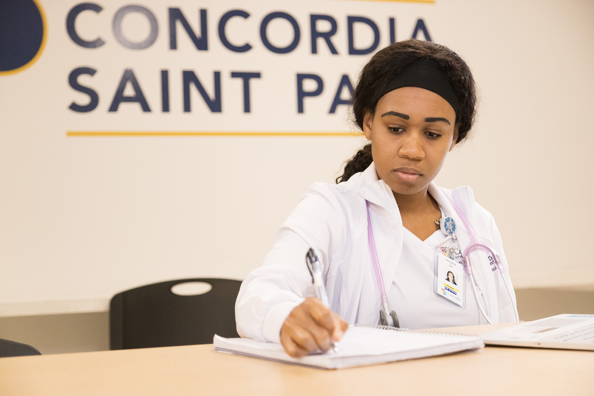 Nursing student wearing white scrubs and a pink stethoscrope sitting at a table writing in a notebook in front of a tan wall with a blue Concordia University, St. Paul logo on it.