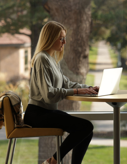 Online student sitting at high table and using a laptop on front of a large window.