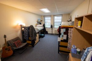 Long angled view of a dorm room with two high bunk beds on either side and two windows at the end of the room.